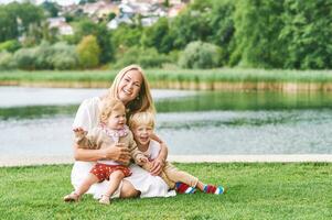 draussen Porträt von glücklich jung Mutter mit zwei schön Kinder spielen durch See oder Fluss auf ein nett Sommer- Tag foto