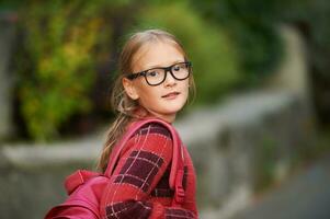 draussen Porträt von bezaubernd wenig Mädchen Gehen zu Schule, waren Rucksack und Brille foto