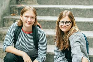 draussen Porträt von zwei Teenager Mädchen Sitzung auf Stufen, tragen zurück zurück, suchen beim das Kamera foto