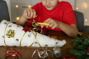 ein süß Junge macht Weihnachten Kunsthandwerk sich selbst. ein Junge schneidet ein Band zum Dekoration mit Schere. handgemacht arbeiten. DIY foto