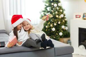 zwei wenig Mädchen im das Santa Hut öffnen Weihnachten Geschenk unter das Weihnachten Baum foto