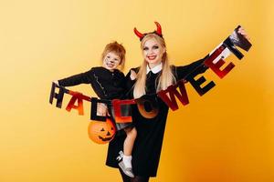 Teufelsfrau, die vor einem gelben Hintergrund mit einem kleinen Mädchen steht und eine Postkarte halloween hält foto