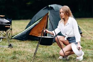 junge Frau, die auf dem Campingplatz kocht, sitzt neben dem Lagerfeuer foto