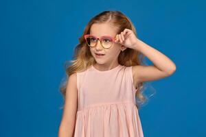 modisch wenig Mädchen im ein Rosa Kleid und Brille ist Stehen auf ein Blau Hintergrund. foto
