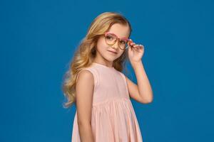 modisch wenig Mädchen im ein Rosa Kleid und Brille ist Stehen auf ein Blau Hintergrund. foto