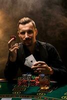 Mann ist spielen Poker mit ein Zigarre und ein Whiskey, ein Mann Show zwei Karten im das Hand, gewinnen alle das Chips auf das Tabelle mit dick Zigarette Rauch. foto