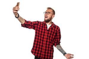 Lebensstil Konzept ein jung Mann mit ein Bart im Hemd halten Handy, Mobiltelefon Telefon und Herstellung Foto von selbst während Stehen gegen Weiß Hintergrund.
