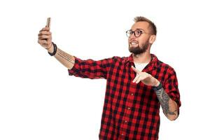Lebensstil Konzept ein jung Mann mit ein Bart im Hemd halten Handy, Mobiltelefon Telefon und Herstellung Foto von selbst während Stehen gegen Weiß Hintergrund.