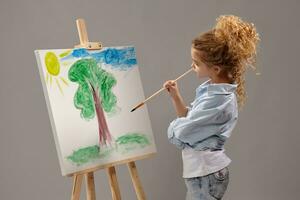 charmant Schule Mädchen ist Gemälde mit ein Aquarell Bürste auf ein Staffelei, Stehen auf ein grau Hintergrund. foto