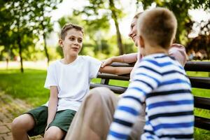 glücklich Mutter ist Sitzung mit ihr Söhne auf Bank im Park. Sie sind reden und genießen ihr Zeit zusammen. foto