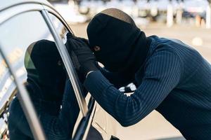 Mann in Schwarz gekleidet mit einer Sturmhaube auf dem Kopf, der vor dem Diebstahl auf das Autoglas schaut. Autodieb, Autodiebstahlkonzept foto