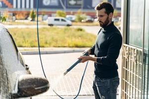 Horizontaler Schuss eines kaukasischen Mannes mit Hochdrucksprüher mit Reinigungsschaum an seinem schwarzen Auto bei Autowaschanlagen im Freien. Sauberes Auto Outdoor-Konzept foto