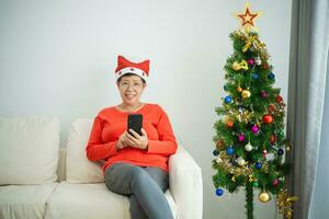 Weihnachten, Weihnachten, Winter Konzept. asiatisch Senior alt Frau mit Handy, Mobiltelefon Telefon zu Plaudern Botschaft lächelnd glücklich heiter. feiert das glücklich Neu Jahr in der Nähe von das Weihnachten Baum im ein Leben Zimmer beim heim. foto