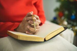 schließen oben Hand. alt Senior Frau tun Hände zusammen und halten Halskette von Kreuze Gebet zu Gott entlang mit das Bibel im das Christian Konzept von Glaube, Spiritualität und Religion. Weihnachten Baum. foto