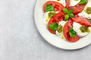 traditionell Italienisch Caprese Salat mit geschnitten Tomaten, Mozzarella, Basilikum, Pesto Soße und würzen auf ein Licht grau Beton Hintergrund. oben Sicht. Kopieren Raum. foto