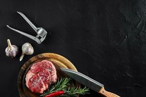 Stück von Rindfleisch Filet, mit Messer zum Schneiden und Hacken Fleisch, Gewürze wurden Kochen - - Rosmarin, Pfeffer, Salz, Knoblauch. auf alt hölzern Tafel auf Stein schwarz Tisch. foto