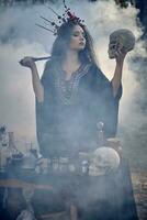 Hexe im Schwarz, lange Kleid, mit rot Krone im ihr Haar. posieren im Kiefer Wald, halten Schädel, Herstellung Trank. Zauber und Hexerei. Nahaufnahme, Rauch. foto