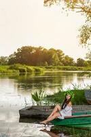 jung Frau mit Blume Kranz auf ihr Kopf, entspannend auf Boot auf Fluss beim Sonnenuntergang. Konzept von weiblich Schönheit, sich ausruhen im das Dorf foto