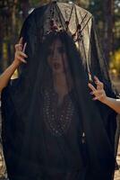 Hexe im Schwarz, lange Kleid, mit rot Krone im ihr lang, lockig Haar unter ein schwarz Schleier. posieren im Kiefer Wald. Zauber, Magie und Hexerei. Nahansicht. foto