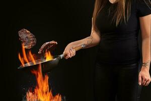 Koch mit tätowiert Hände, gekleidet im Gamaschen und T-Shirt. halten Wok schwenken über Feuer und braten zwei Rindfleisch Steaks gegen schwarz Hintergrund. Seite Aussicht foto