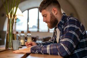 jung bärtig Geschäftsmann sitzt im Cafe, Zuhause beim Tabelle und schreibt im Notizbuch. Mann ist Arbeiten, studieren. foto