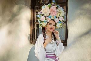 Brünette Mädchen im ein Weiß ukrainisch authentisch National Kostüm und ein Kranz von Blumen ist posieren gegen ein Weiß Hütte. Nahansicht. foto