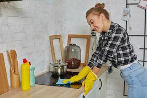 blond Frau im schützend Handschuhe mit Lappen Reinigung elektrisch Herd beim Zuhause Küche. Mädchen Waschen schwarz glänzend Oberfläche von Küche oben foto