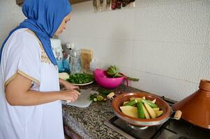 Muslim Hausfrau Frau Hacken Grün Bohnen auf Schneiden Planke, Kochen marokkanisch Tajine Stehen durch Marmor Küche Zähler foto