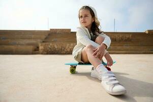 schön Kind Mädchen verträumt suchen weg, Sitzung auf ihr Blau Skateboard mit mehrfarbig Räder, auf das städtisch Skatepark foto