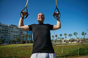 Porträt von ein jung Erwachsene Sportler mit Suspension Riemen, tun Oberer, höher Körper Fitness Übungen draußen foto