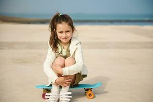 voll Größe Porträt von ein wenig Mädchen im beiläufig Kleidung, Sitzung auf ihr Penny Tafel auf ein Spielplatz auf das Meer Promenade foto