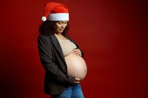 charmant Mitte alt schwanger Frau warnen ein Santa Hut, halten ihr nackt Bauch, lächelnd, isoliert Über rot Hintergrund foto