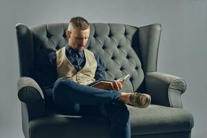 Mann mit stilvoll Schnurrbart, gekleidet im schwarz Hemd und Hose, Beige Weste ist Sitzung auf dunkel Sofa, lesen ein Zeitschrift. grau Hintergrund, Nahansicht. foto