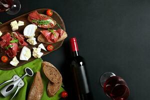 köstlich Vorspeise zu Wein - - Schinken, Käse, Stangenbrot Scheiben, Tomaten, serviert auf ein hölzern Planke, und Glas mit rot Wein auf schwarz Oberfläche foto