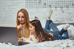 schön jung Mutter und ihr süß Tochter im Weiß Pullover und Jeans Lügen auf das Bett beim heim, Lachen und suchen im Laptop foto