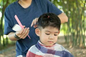 Barbier Schneiden Haar von ein asiatisch Junge im ein öffnen Raum gefüllt mit Bäume. foto