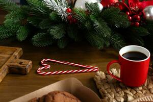 rot Tasse von Kaffee, Süßigkeiten Stöcke geformt wie Herz, und Teller von Kekse auf hölzern Tabelle mit Fichte Geäst auf Hintergrund foto