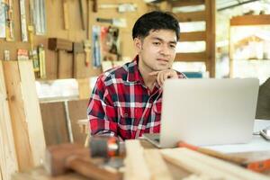 asiatisch Mann Inhaber ein klein Möbel Geschäft ist überprüfen bestellen online. Zimmermann männlich ist einstellen Holz zu das gewünscht Größe. Architekt, Designer, eingebaut, Fachmann Holz, Handwerker, Werkstatt. foto