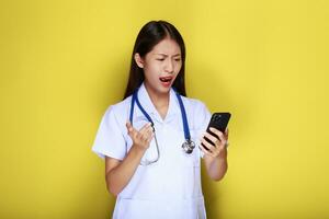 Porträt von ein schön jung Frau im ein Gelb Hintergrund, asiatisch Frau posiert mit ein Zelle Telefon während tragen ein Arzt Uniform und ein Stethoskop. foto