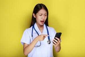 Porträt von ein schön jung Frau im ein Gelb Hintergrund, asiatisch Frau posiert mit ein Zelle Telefon während tragen ein Arzt Uniform und ein Stethoskop. foto