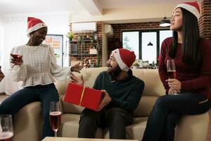 aufgeregt Mann mit festlich eingewickelt Geschenk Box raten Geheimnis Santa während feiern Weihnachten im Büro. heiter Mitarbeiter im Weihnachten Hüte austauschen die Geschenke und genießen Neu Jahr Urlaub foto