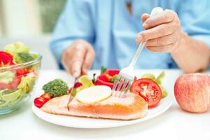 asiatisch Alten Frau geduldig Essen Lachs Anteil und Gemüse Salat zum gesund Essen im Krankenhaus. foto