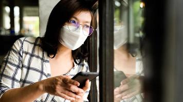 Eine Frau, die eine medizinische Maske trägt und ein Smartphone hält, schaut aus dem Fenster. foto