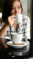 Nahaufnahme erschossen eine weiße Kaffeetasse steht auf dem Tisch, im Hintergrund sitzt eine junge Frau, die Kaffee trinkt. foto