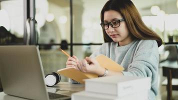 asiatische Studentin mit Brille, die Laptop betrachtet und Notizen in einem Notizbuch in der Hand macht