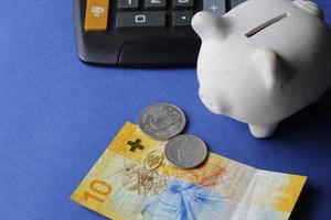 Schweizer Banknote, Münzen, weißes Sparschwein und Taschenrechner auf blauem Hintergrund foto