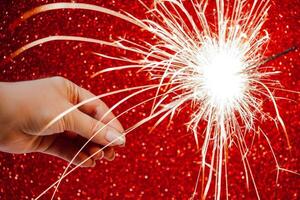 Neu Jahr Feier. Frauen Hand hält hell Wunderkerze auf rot Hintergrund. Weihnachten Nacht. hell Funken von Feuer und Bokeh. foto