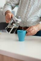im Ruhestand Mann halten Blau Tasse und Französisch Drücken Sie auf Küche Tabelle Gießen heiß aromatisch trinken von ein Kaffee Maschine während Frühstück. Alten Person im das Morgen Trinken frisch braun Espresso von Becher foto