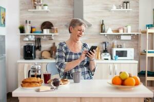 Single Senior Frau Surfen auf Telefon im Küche während Frühstück. authentisch Alten Person mit modern Smartphone Internet Technologie genießen Freizeit Zeit mit Gadget beim Pensionierung Alter foto