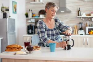 glücklich Senior Frau schieben auf Französisch Drücken Sie während vorbereiten Kaffee zum Frühstück. Alten Person im das Morgen genießen frisch braun Cafe Espresso Tasse Koffein von Jahrgang Becher Filter entspannen Erfrischung foto